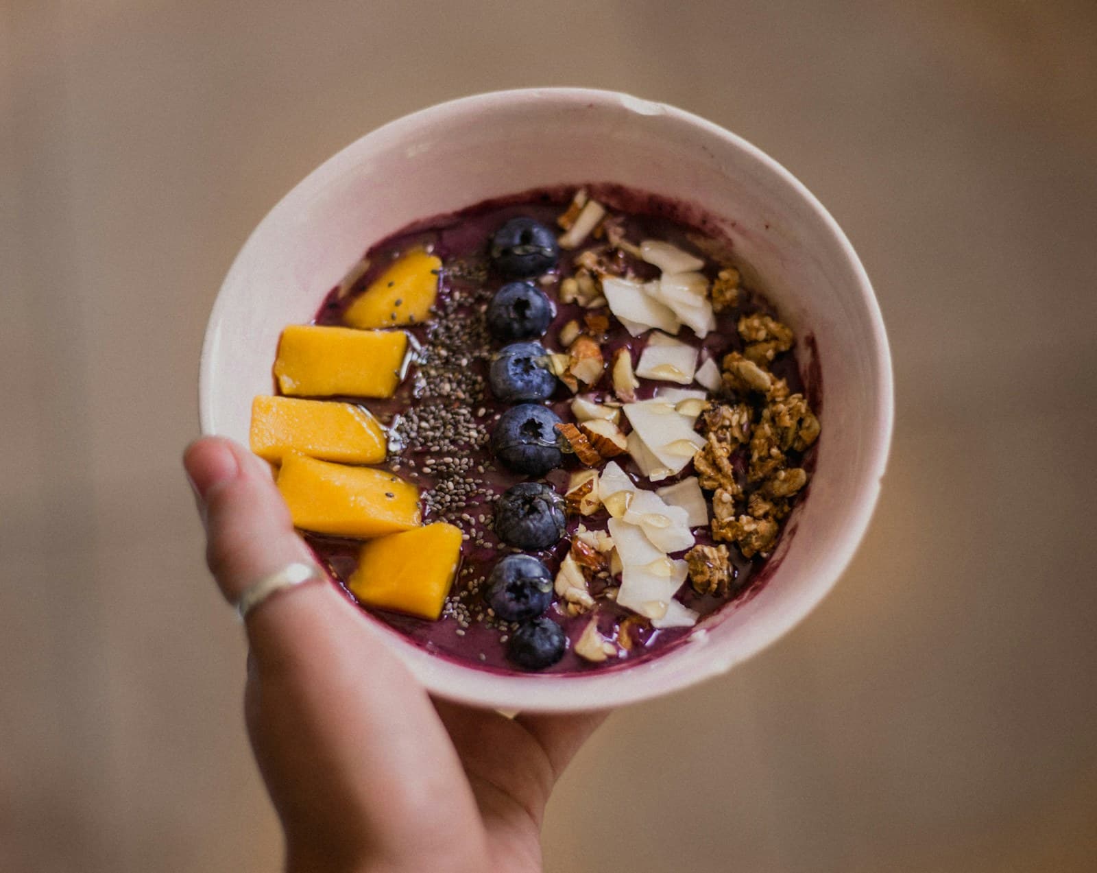Hand holding an acai bowl topped with mango, blueberries, granola, chia, and coconut