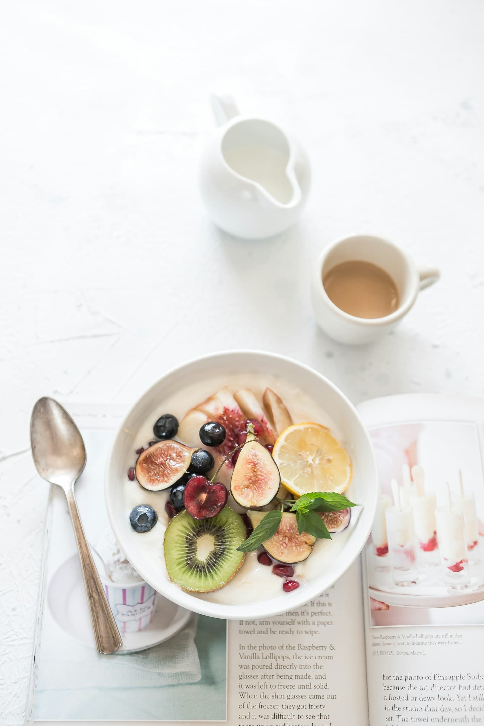 Yogurt bowl with figs, kiwi, cherries, blueberries, and lemon beside a cup of coffee