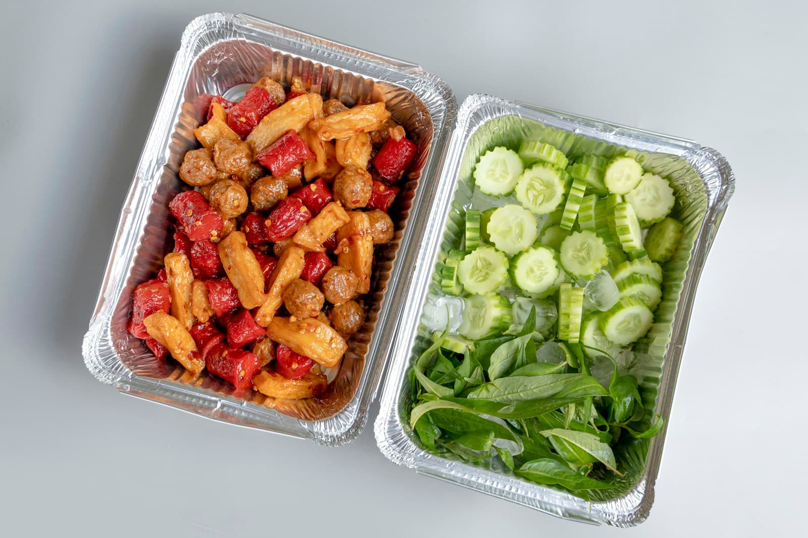 Two meal-prep trays with cooked protein, peppers, cucumber, and fresh herbs