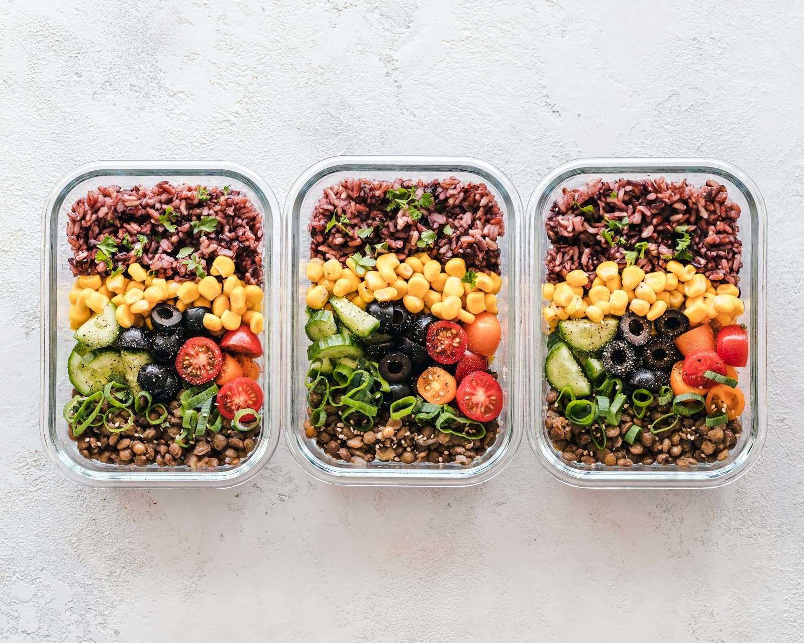 Three meal-prep containers with grains, lentils, corn, olives, cucumber, and tomatoes